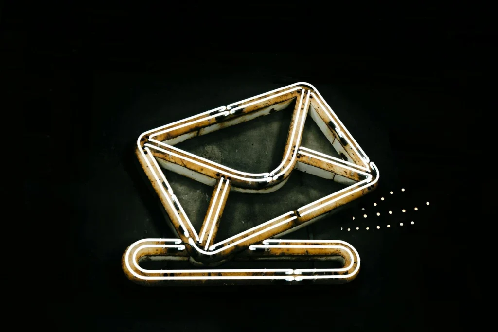 A glowing neon sign on a black background depicts an envelope above a paperclip, symbolizing email or messaging. The sign is outlined in white and yellow lights, giving it a modern, minimalistic look.