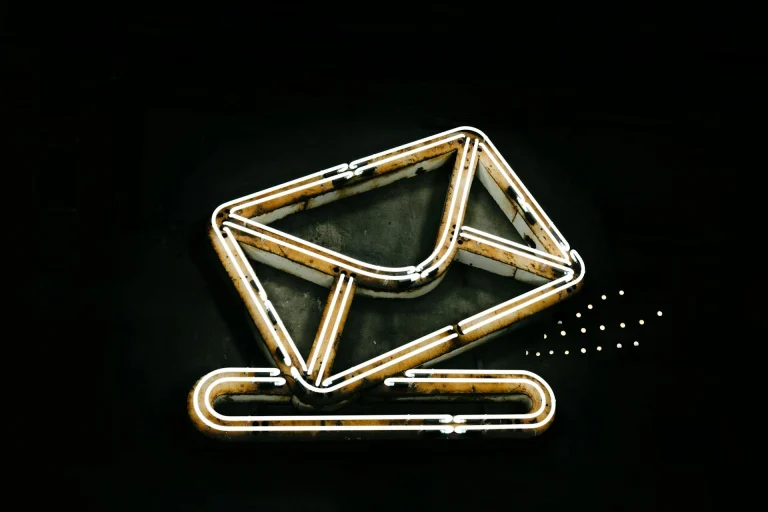 A glowing neon sign on a black background depicts an envelope above a paperclip, symbolizing email or messaging. The sign is outlined in white and yellow lights, giving it a modern, minimalistic look.