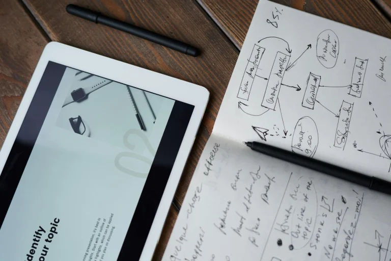 A tablet displaying a website lies on a wooden table next to a notepad with handwritten notes and sketches. A black pen and stylus are also on the table.