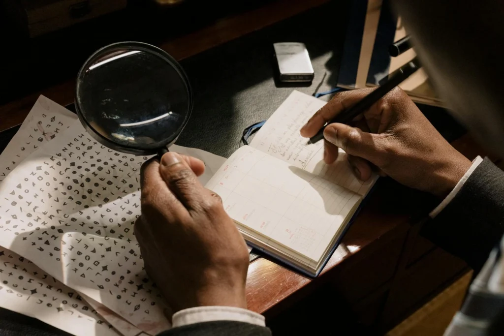 A person holds a magnifying glass over a sheet with symbols and writes notes in an open notebook on a desk, suggesting they are solving a code or puzzle.