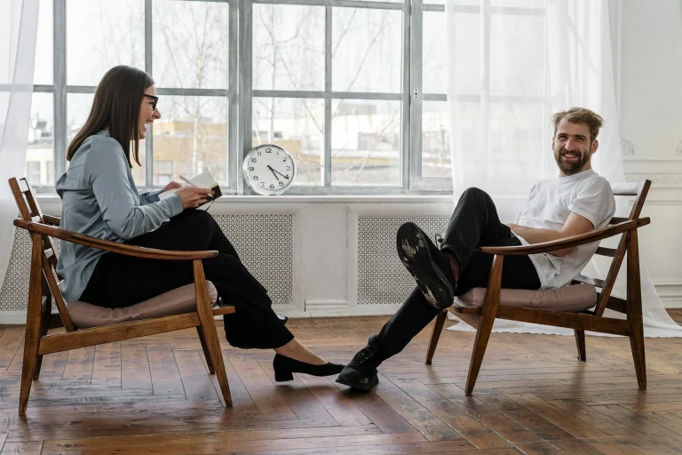 A woman and a man sit facing each other in wooden chairs in a bright room. The woman holds a notebook and pen, while the man sits casually, smiling. A clock sits on the windowsill behind them.