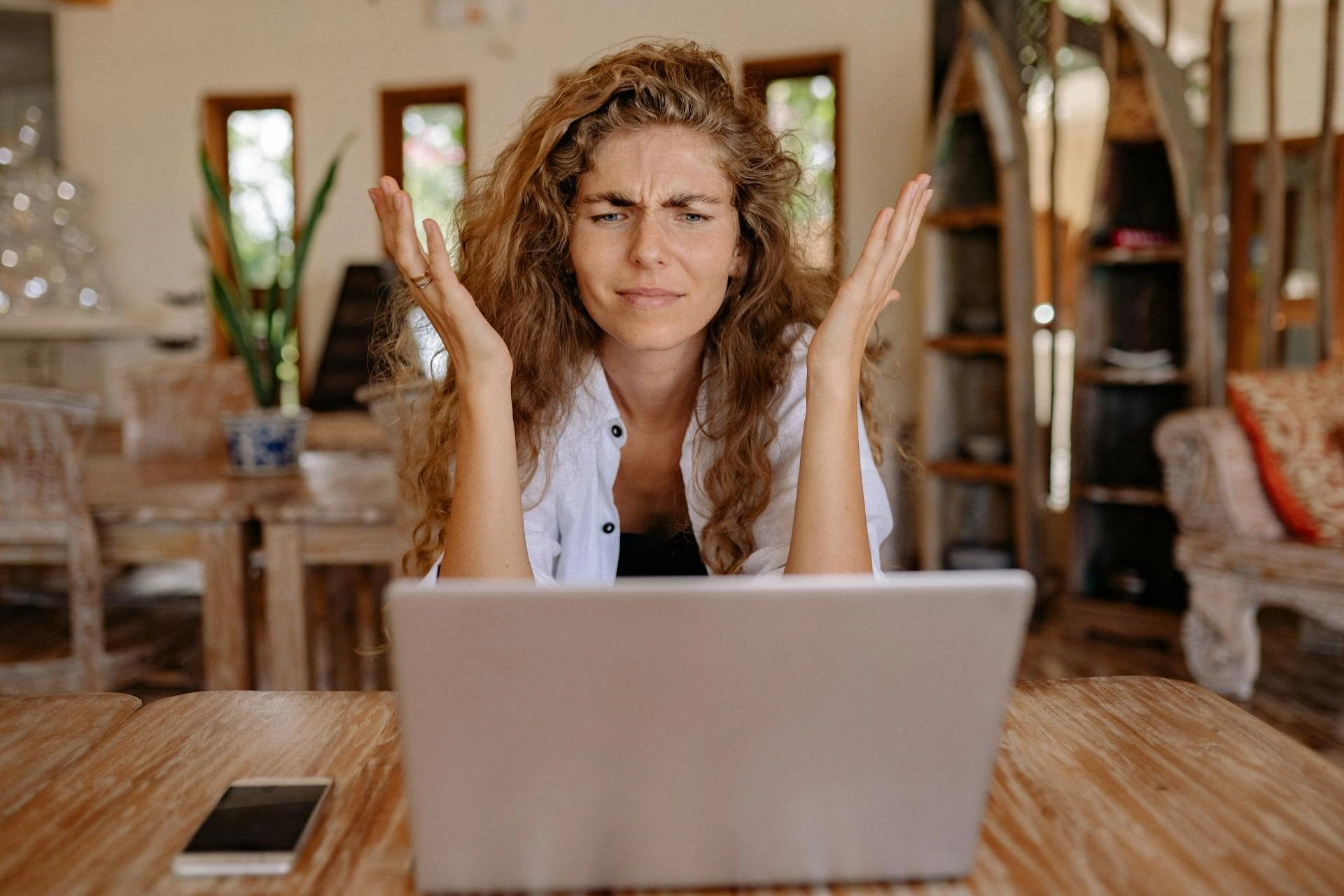 A woman with curly hair sits at a table in front of a laptop, raising her hands in frustration and frowning. A smartphone is on the table beside her. The setting appears to be a cozy, wooden interior.