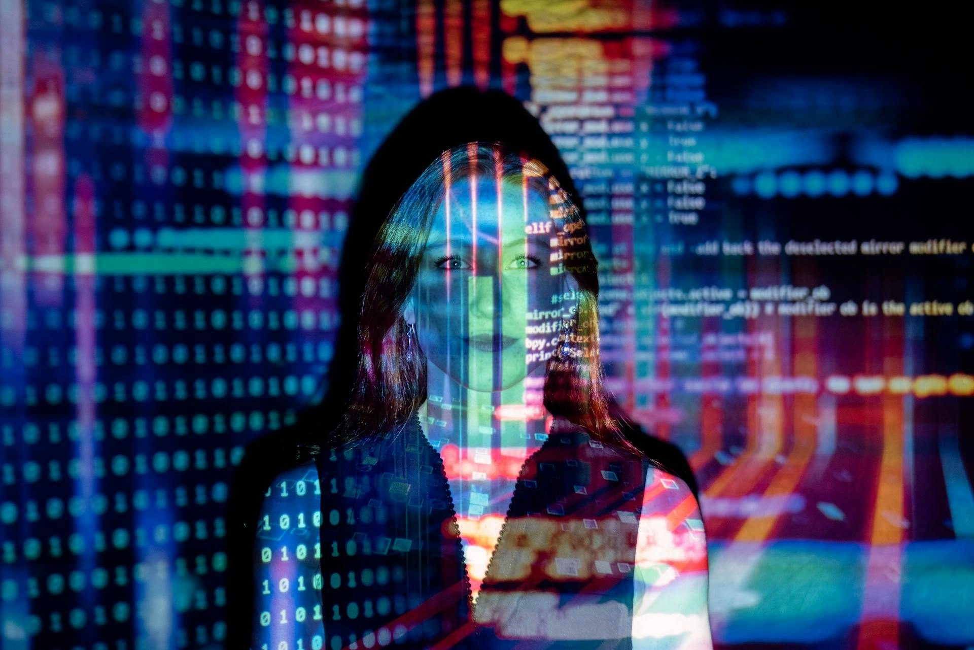 A person stands in front of a screen displaying colorful binary code, programming text, and circuit patterns, with the digital graphics projected onto their face and body.
