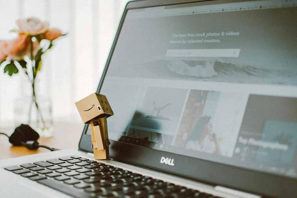 A small cardboard robot figure stands on the keyboard of an open Dell laptop, which displays a website with stock photos. A blurred vase with pink flowers is in the background.