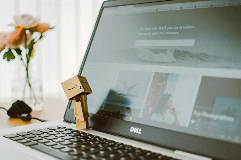 A small cardboard robot figure stands on the keyboard of an open Dell laptop, which displays a website with stock photos. A blurred vase with pink flowers is in the background.
