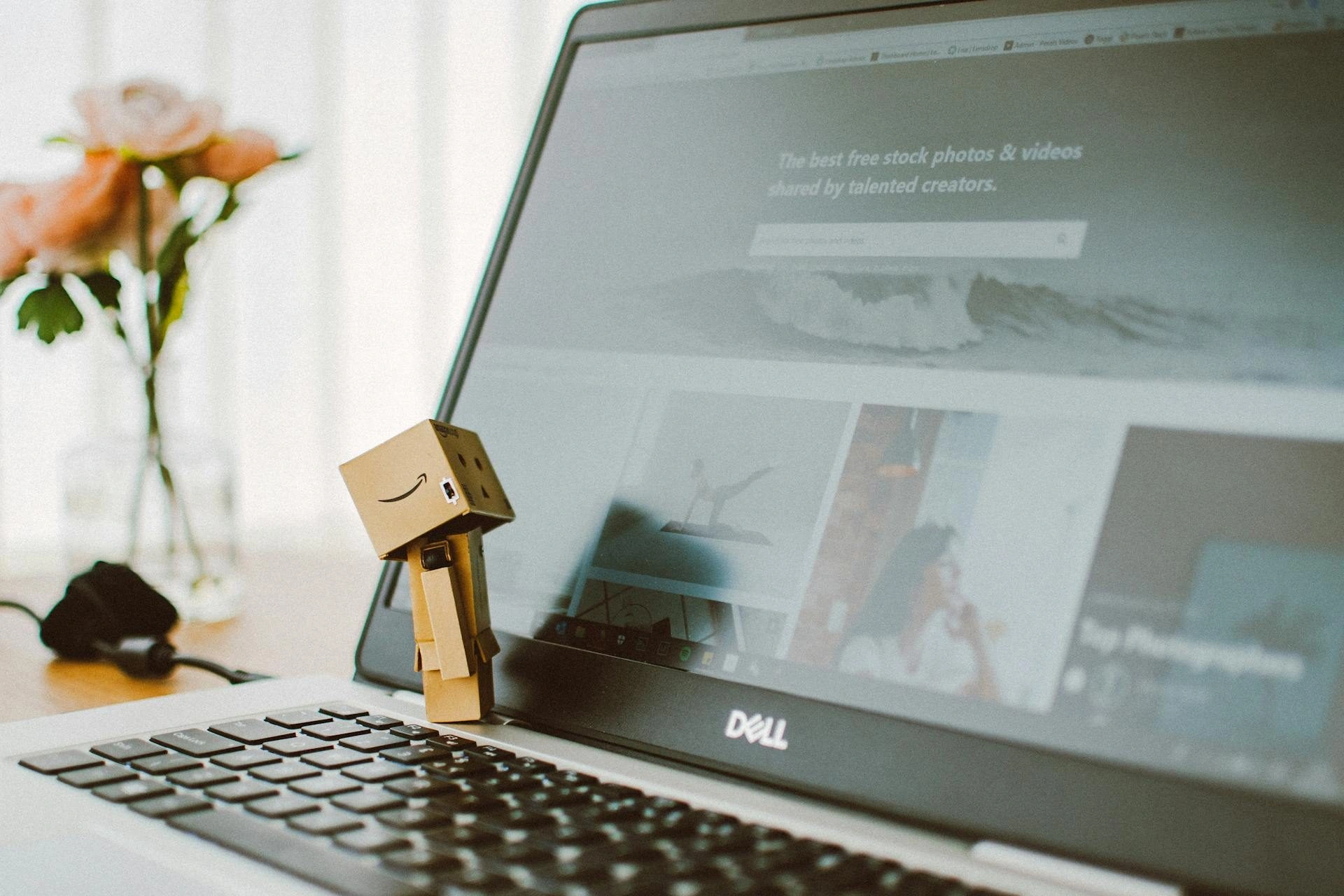 A small cardboard robot figure stands on the keyboard of an open Dell laptop, which displays a website with stock photos. A blurred vase with pink flowers is in the background.