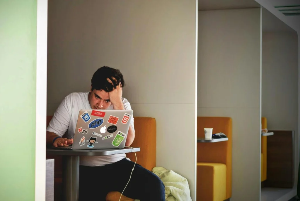 A person sits alone in a booth, looking stressed with their hand on their head, working on a laptop covered in tech-related stickers. A takeaway cup sits on the table beside them.