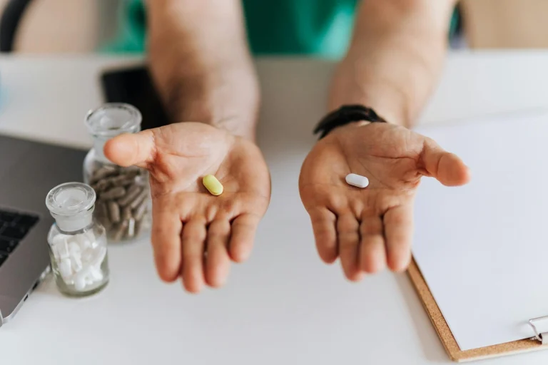 A person holds a yellow pill in one hand and a white pill in the other, seated at a desk with jars of capsules, a laptop, and a clipboard in view.