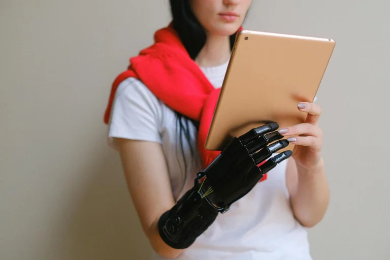 A woman with long dark hair, wearing a white shirt and red sweater, holds a tablet with a black prosthetic hand against a plain light background.