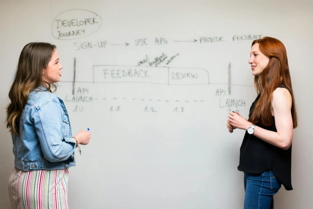 Two women stand in front of a whiteboard discussing a diagram labeled “Developer Journey,” which includes stages like sign-up, API launch, feedback, and review. Both are engaged in conversation.