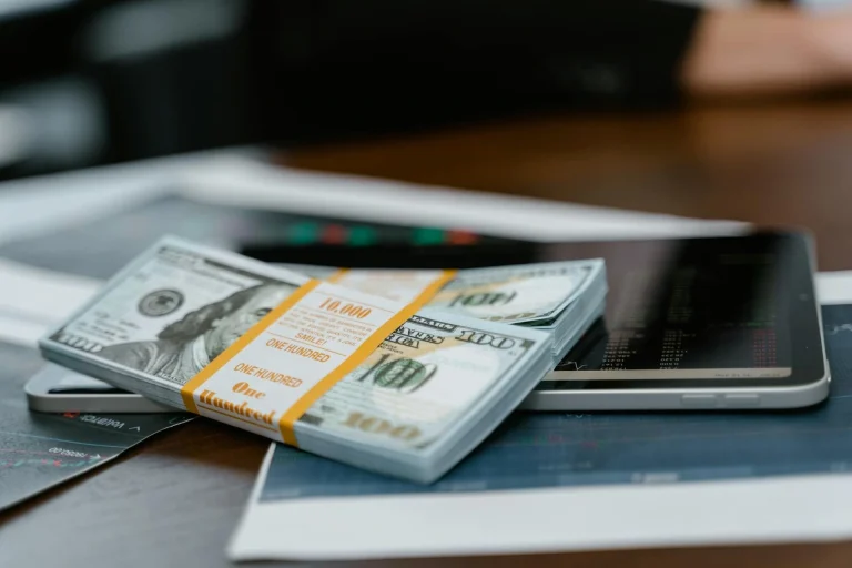 A stack of one hundred dollar bills wrapped in a band sits on top of a tablet, with financial charts and papers scattered on a desk in the background.