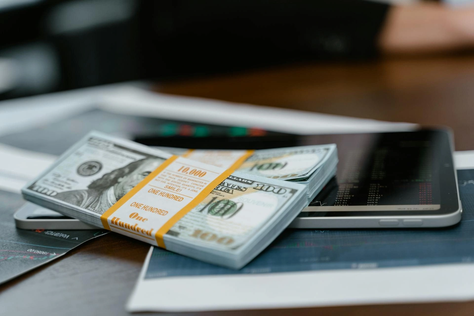 A stack of one hundred dollar bills wrapped in a band sits on top of a tablet, with financial charts and papers scattered on a desk in the background.