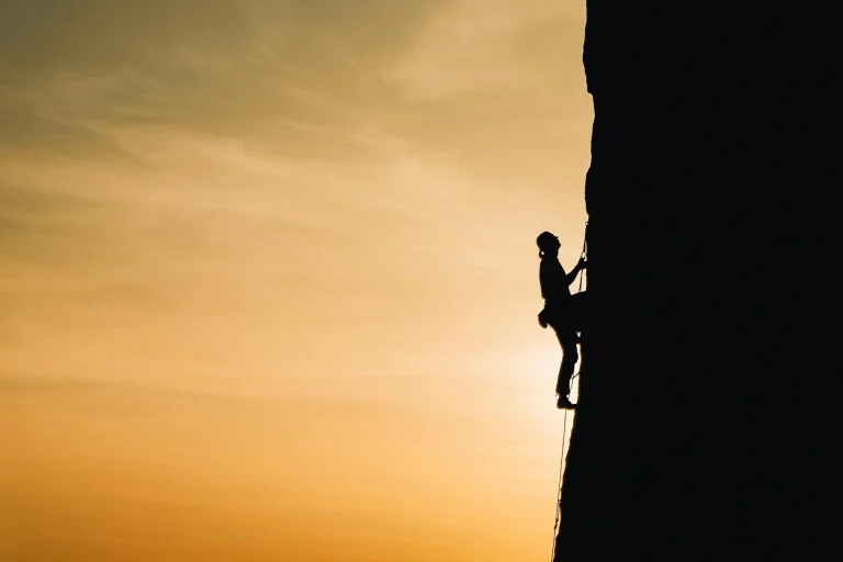 A person climbs a steep rock face, silhouetted against a golden sunset sky, highlighting the challenge and adventure of outdoor rock climbing.