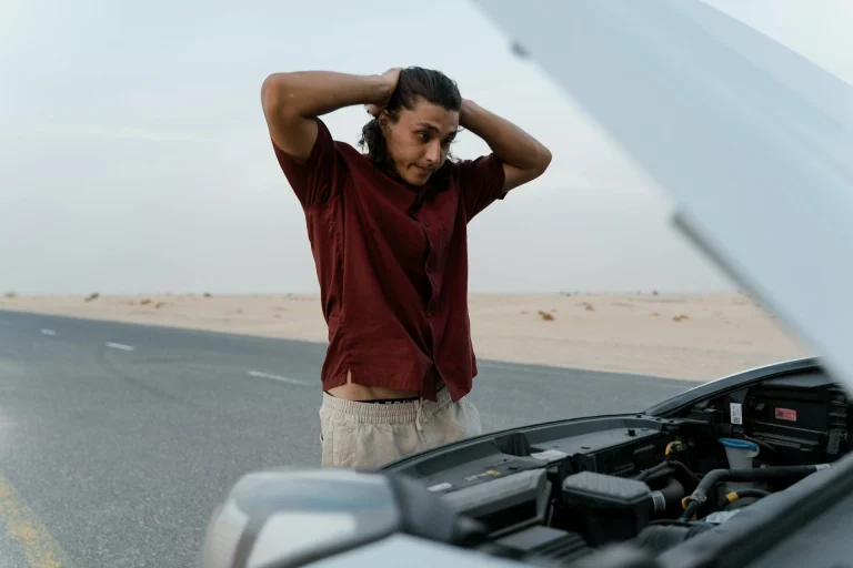 A person stands by the side of a desert road, looking worried with their hands on their head, while inspecting the engine of a car with the hood open.