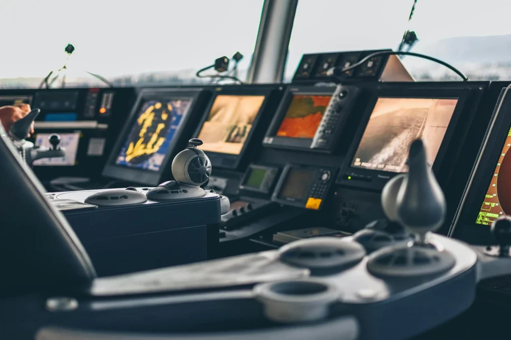 Close-up view of a modern ship’s control room with multiple digital navigation screens, control panels, and levers, showing advanced maritime technology and equipment.
