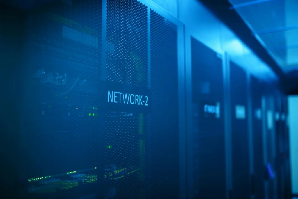 Close-up of a server room with blue lighting, showing server racks labeled "NETWORK-2" and faint green indicator lights inside the equipment.