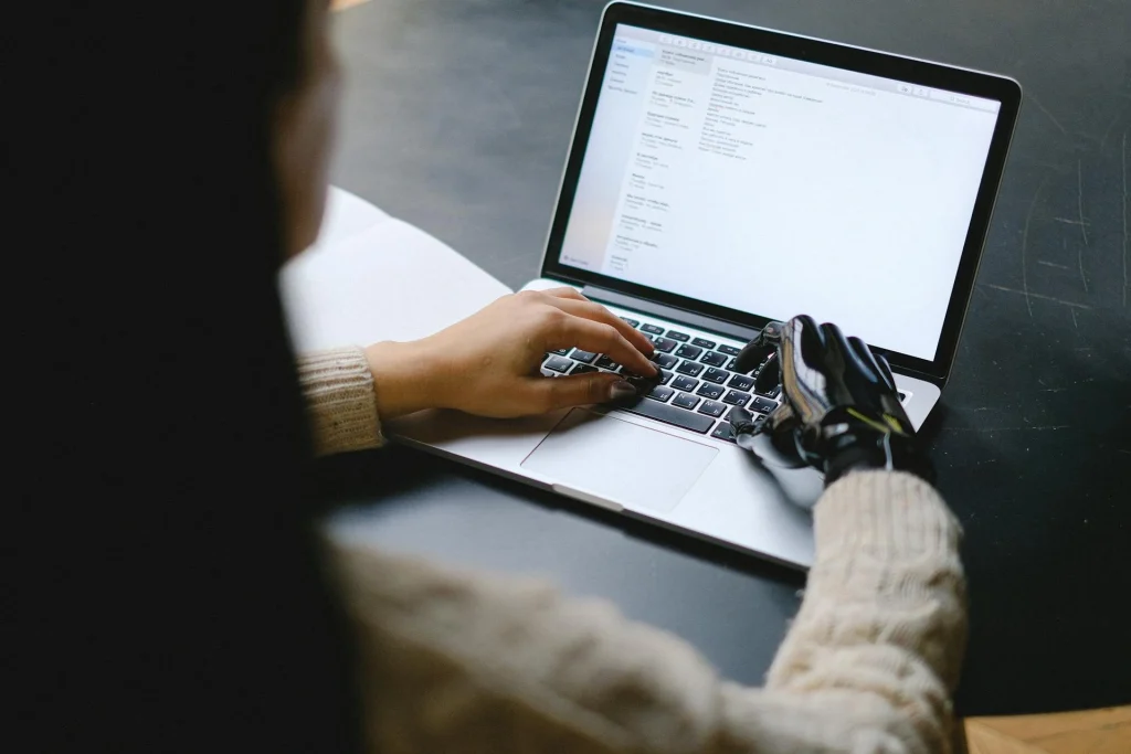 A person wearing a light sweater and a black prosthetic hand types on a laptop at a dark desk, with text displayed on the laptop screen.