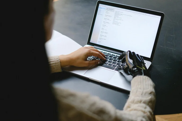 A person wearing a light sweater and a black prosthetic hand types on a laptop at a dark desk, with text displayed on the laptop screen.