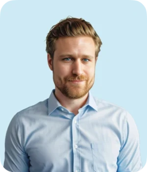 A man with light brown hair and a beard, wearing a light blue button-up shirt, stands in front of a plain light blue background, looking ahead with a slight smile.