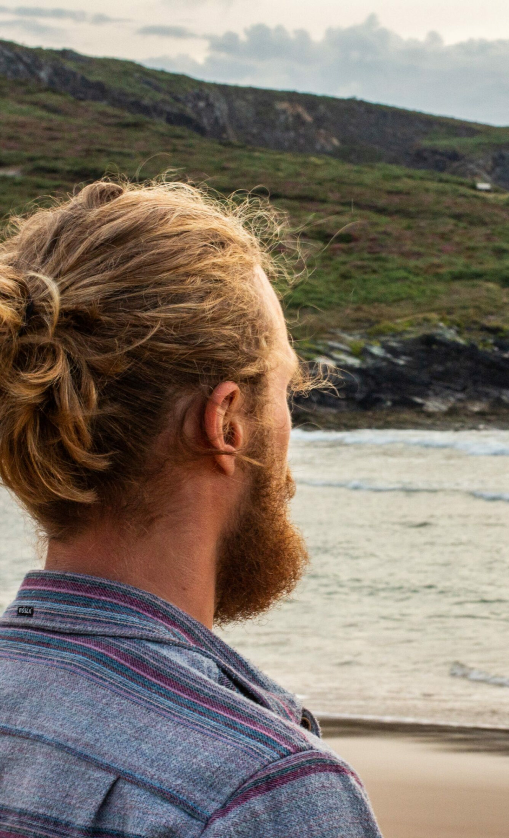 Una persona con el pelo rubio rizado recogido en un moño y barba está de pie en una playa, frente al océano. Al fondo se ven colinas cubiertas de vegetación bajo un cielo nublado.