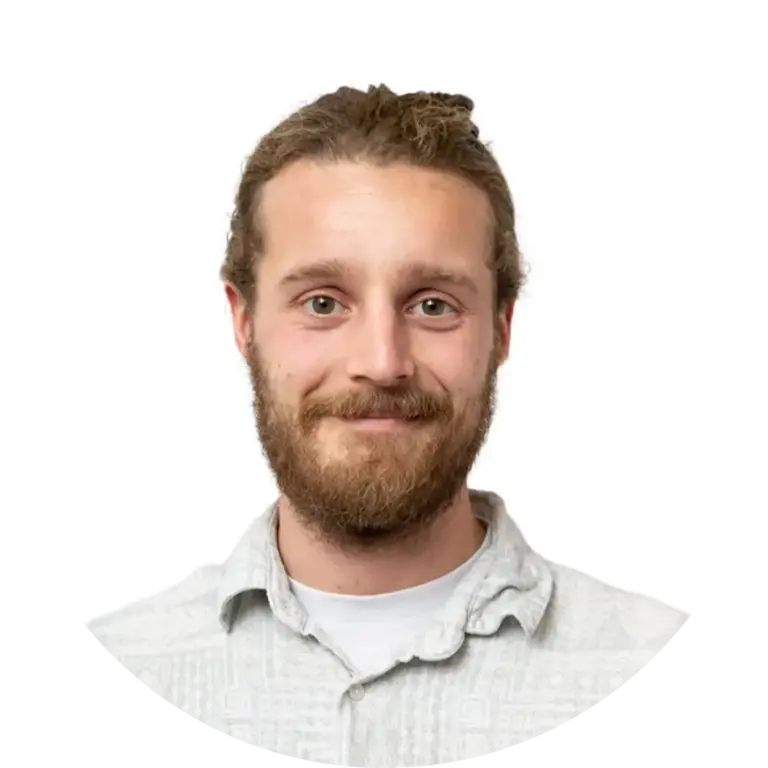 Ein junger Mann mit heller Haut, einem braunen Bart und langen, zurückgebundenen Haaren lächelt in die Kamera. Er trägt ein helles, gemustertes Button-up-Hemd. Der Hintergrund ist einfarbig weiß und hat einen runden Rahmen.