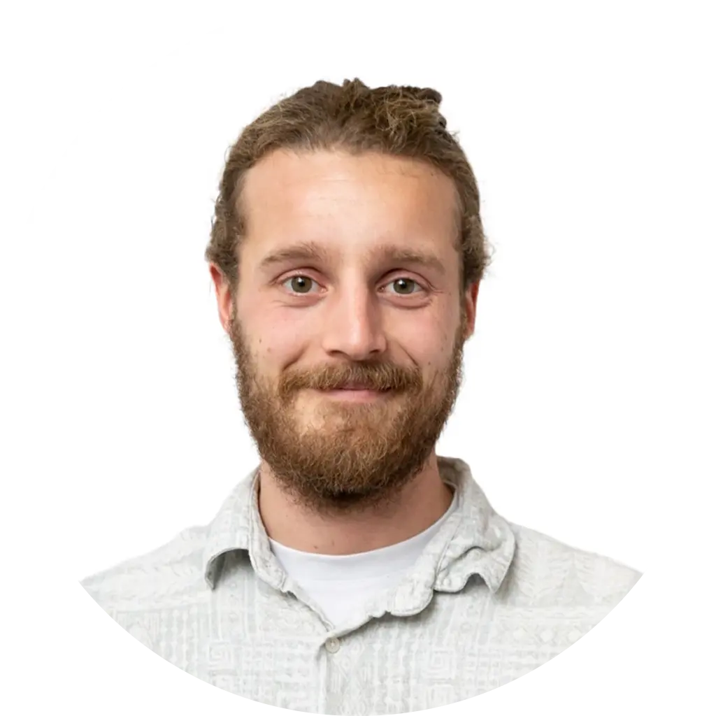 A young man with light skin, a brown beard, and long hair tied back smiles at the camera. He is wearing a light-colored, patterned button-up shirt. The background is plain white in a circular frame.
