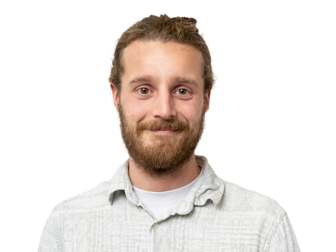 A man with light brown hair and a beard, wearing a light gray patterned button-up shirt, stands facing the camera and smiles softly. The background is plain black.