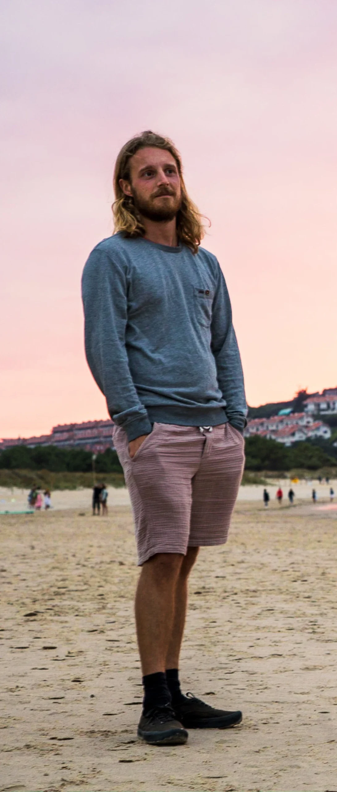A man with long blond hair, wearing a gray long-sleeve shirt, pink shorts, black socks, and black shoes stands on a sandy beach in Suances, Cantabria at sunset, with buildings and trees in the background.