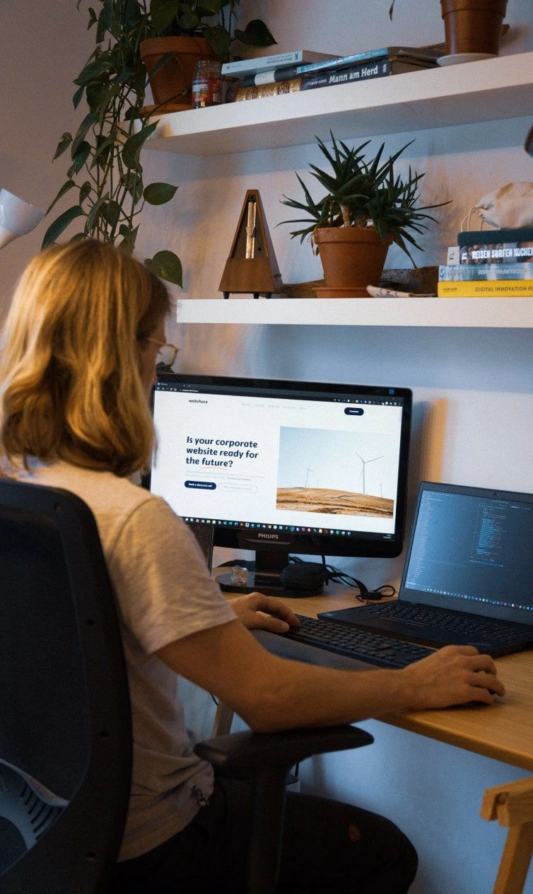 A person with long blond hair works at a desk with a monitor, laptop, and keyboard. The monitor displays a website, and shelves with potted plants and books are visible in the background.