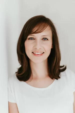 A woman with straight brown hair and blue eyes is smiling softly. She is wearing a white shirt and standing against a plain, light-colored background. The lighting is soft and natural.
