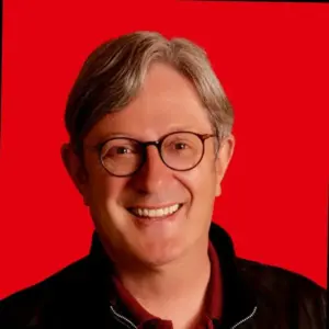 A smiling person with gray hair and glasses, wearing a dark jacket and a maroon shirt, in front of a solid bright red background.
