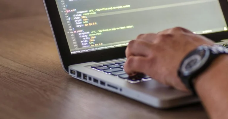A close-up of a person’s hand typing on a laptop keyboard with code visible on the screen, including CSS styling rules for layout and background images.