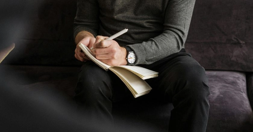 A person in a gray sweater and black pants sits on a dark couch, writing in a notebook with a pen. Only their hands, notebook, and part of their torso are visible.