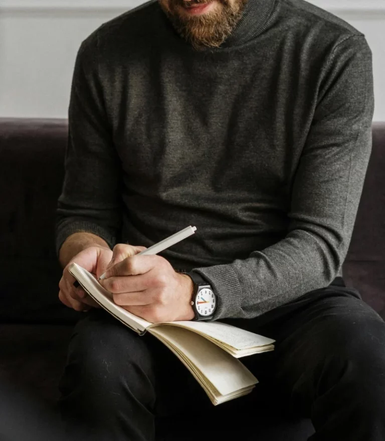 A person in a gray sweater and black pants sits on a dark couch, writing in a notebook with a pen. Only their hands, notebook, and part of their torso are visible.