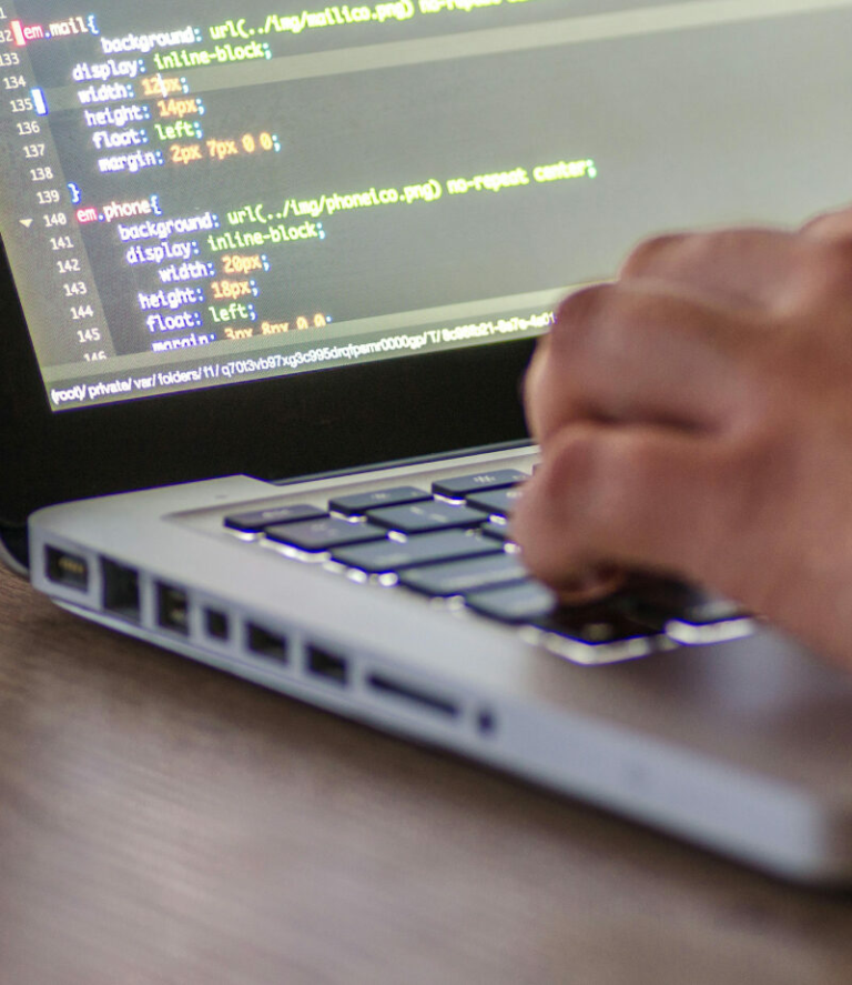 A close-up of a person’s hand typing on a laptop keyboard with code visible on the screen, including CSS styling rules for layout and background images.