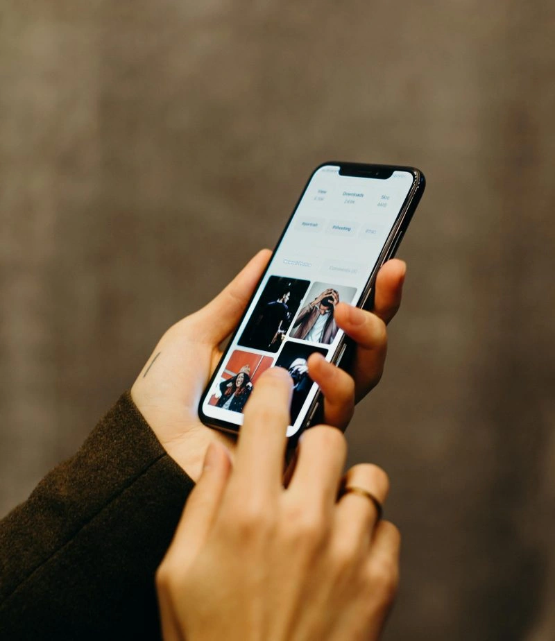 A person holding a smartphone and browsing a photo gallery app, with their thumb about to tap on an image. The background is blurred and the person is wearing a dark sleeve and a ring.