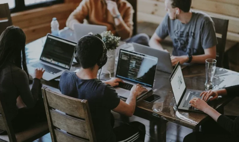 Five people sit around a wooden table, each working on laptops. Some wear headphones and are focused on their screens. The environment appears casual, collaborative, and modern, possibly in a coworking space or office.