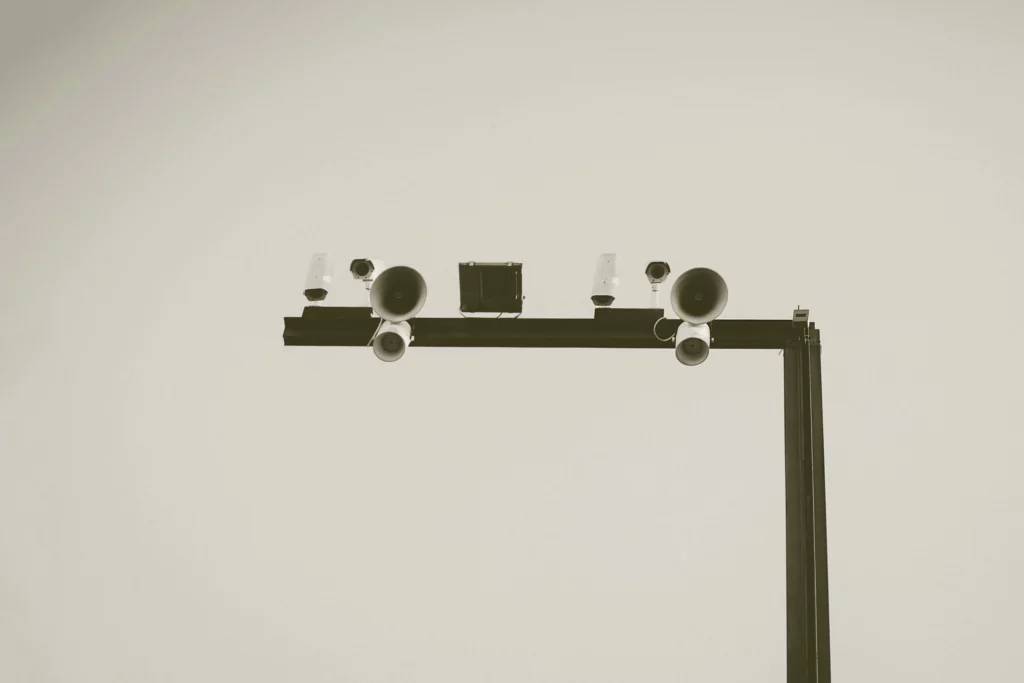 A black metal pole holds speakers, cameras, and lights against a plain, light sky. The image is minimalistic and monochrome, emphasizing the structure and equipment.