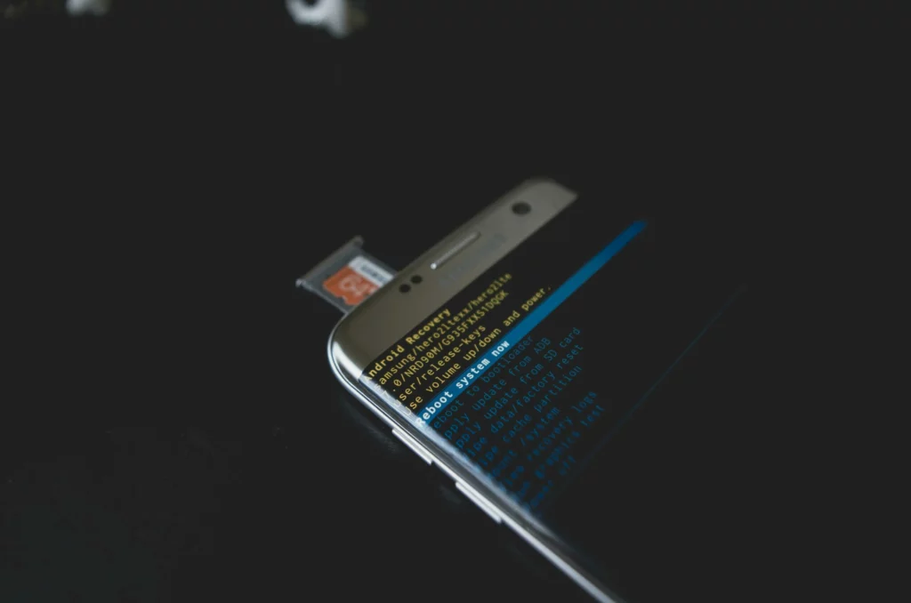 A smartphone with a microSD card partially inserted displays a recovery mode screen with yellow and blue text on a black background. The phone is on a dark surface with a black background.