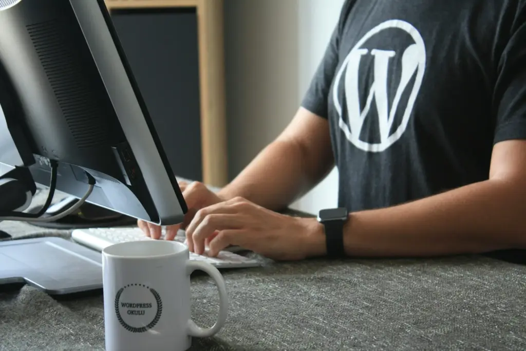 Jemand in einem WordPress-T-Shirt tippt an einem Schreibtisch mit Computerbildschirm auf einer Tastatur. Im Vordergrund steht eine weiße Tasse mit der Aufschrift "WordPress Oku".